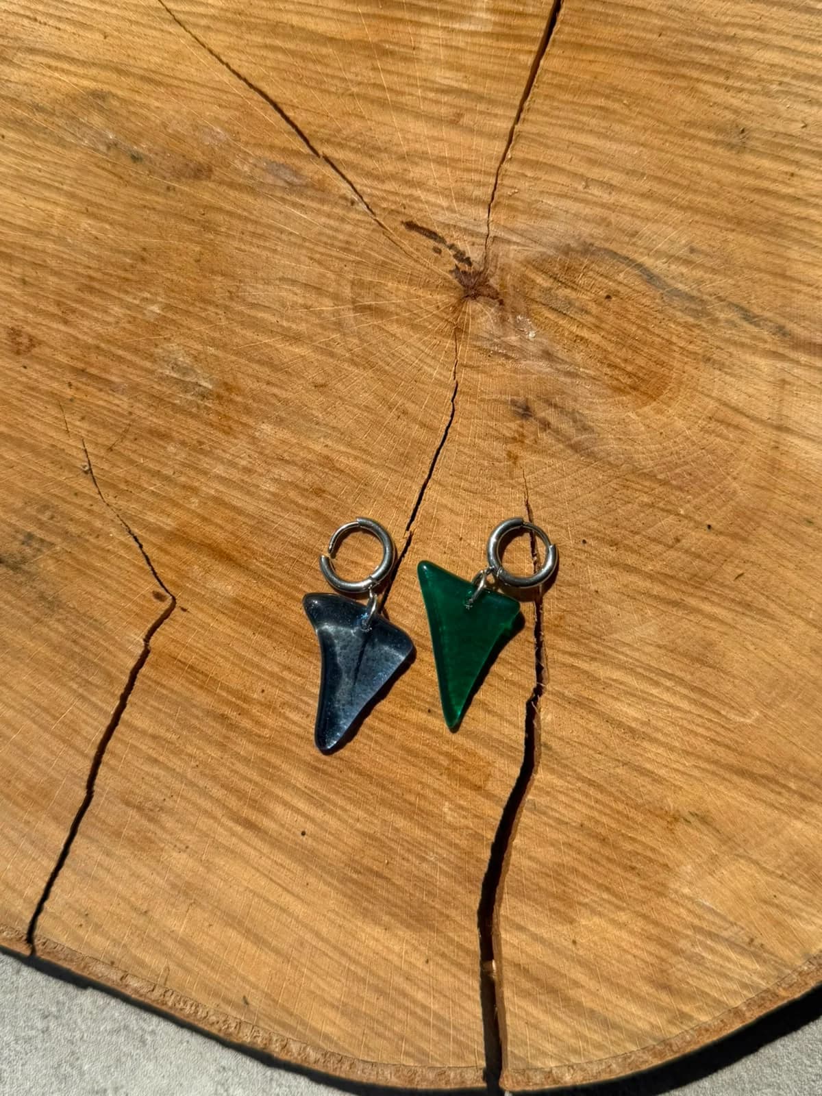 Main tenant deux boucles d'oreilles triangulaires en verre bleu translucide et vert près d'une plante.