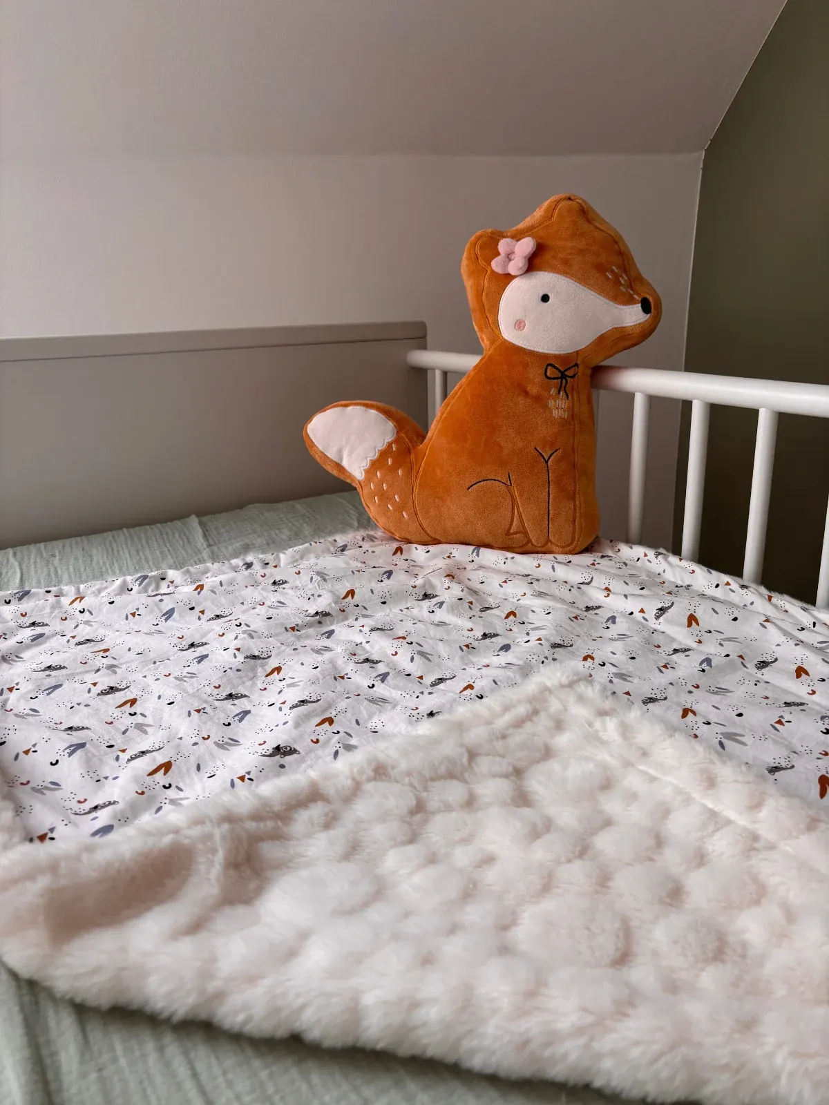 Couverture bébé imprimée et doublée de polaire étalée sur un lit d'enfant à côté d'une peluche renard.