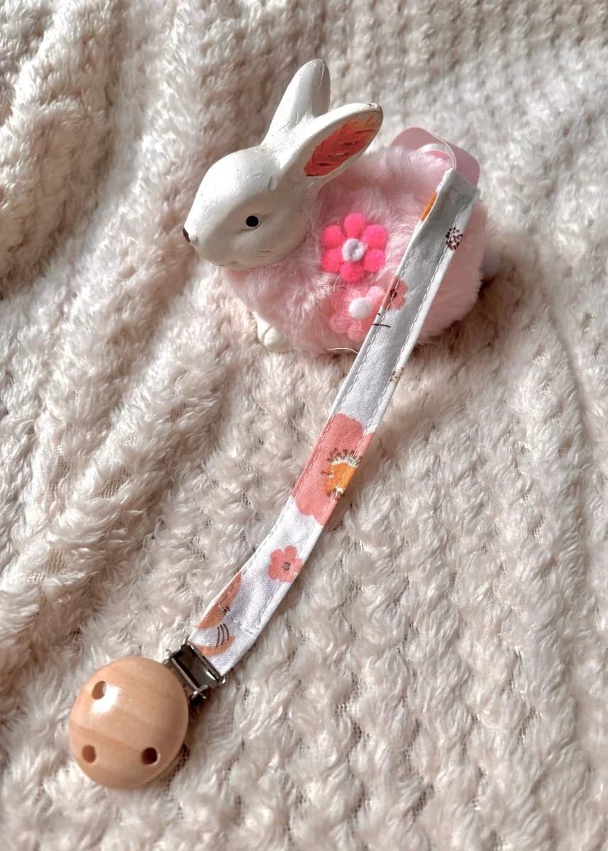 Attache-tétine en tissu à fleurs avec clip rond en bois naturel, posée à côté d'une figurine lapin.