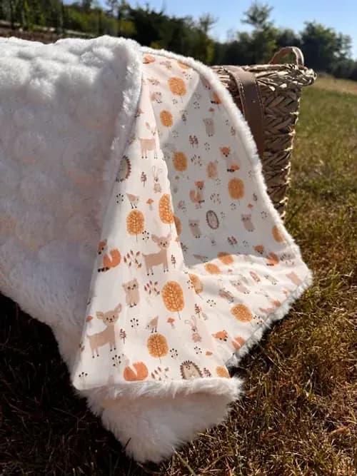 Couverture bébé motif animaux d'automne et revers polaire blanc sur un lit à côté d'un coussin renard.