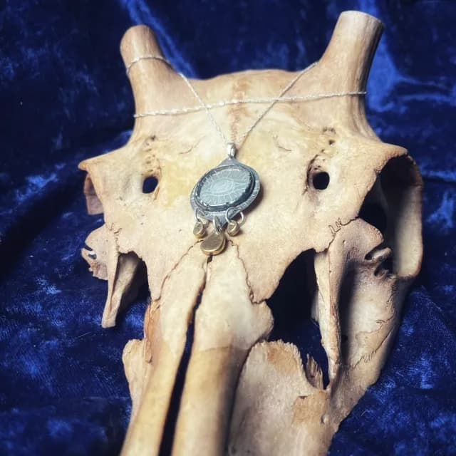 Pendentif œil en argent et laiton présenté sur une chaîne fine posée sur un crâne.